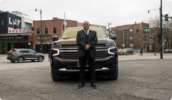 A man in a black suit stands confidently in front of a black SUV on a city street.