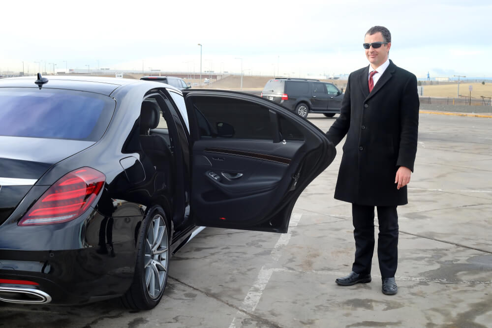 Chauffeur from limo services holding a car door open beside a black luxury sedan at an airport pickup area.
