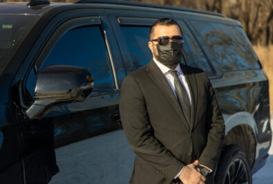 Man in a black suit and mask standing beside a black car.